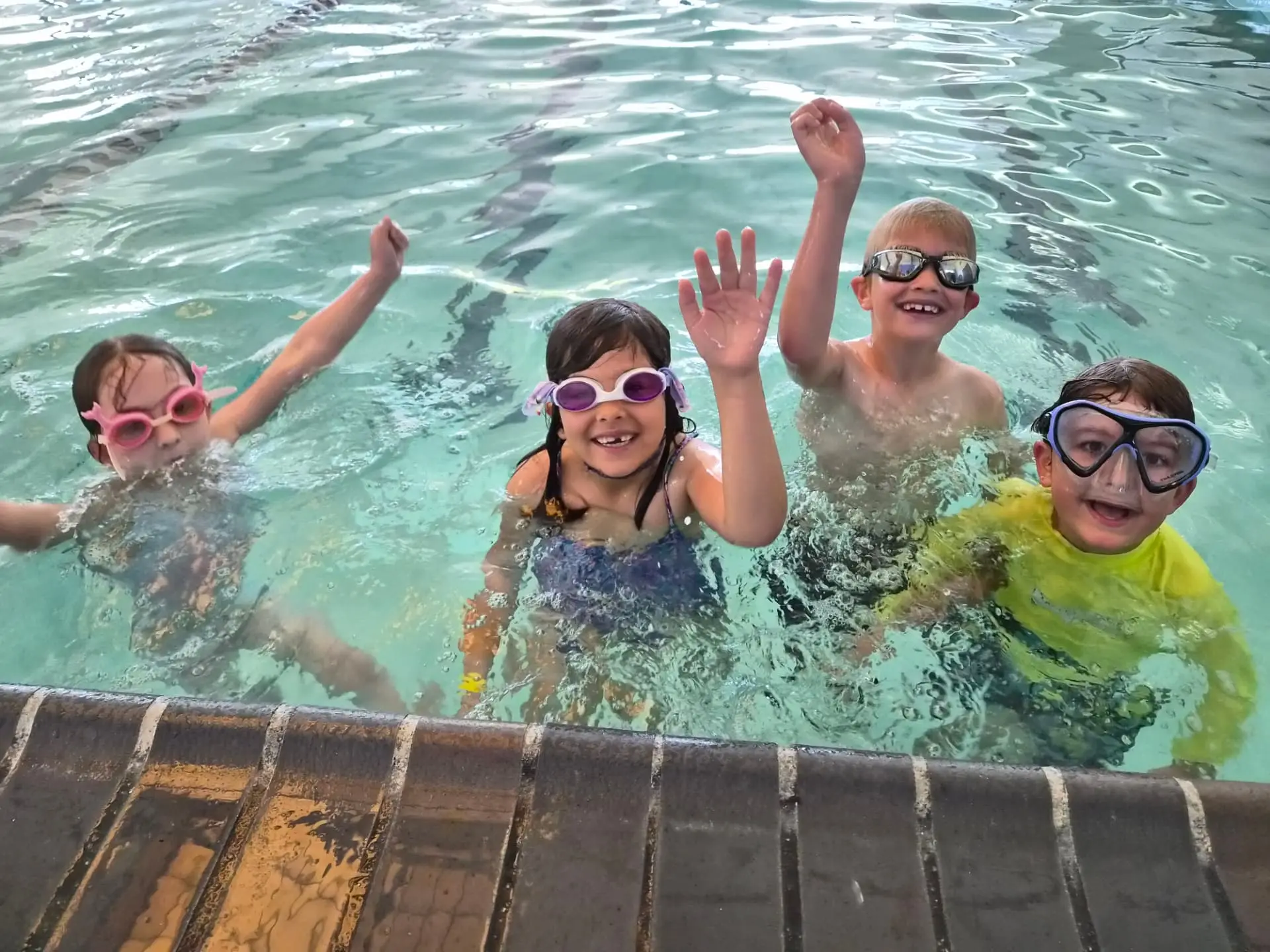 Four kids in goggles waving from the heated indoor pool at a HealthSPORT pool party